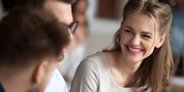 A young woman with long, blonde hair smiles brightly while engaging with two people nearby. The setting appears to be an informal gathering or discussion among friends.