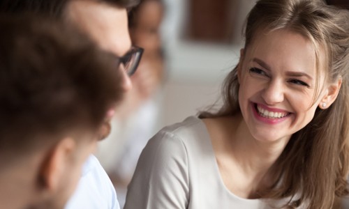 A young woman with long, blonde hair smiles brightly while engaging with two people nearby. The setting appears to be an informal gathering or discussion among friends.