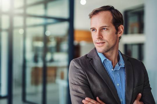 A man in a suit poses thoughtfully with arms crossed, standing in a modern, softly lit office with glass walls and a blurred background of workspace elements.