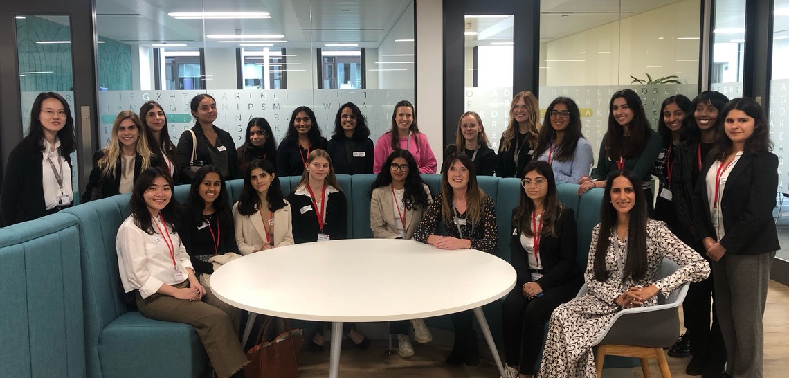 A group of diverse women poses together around a circular table in a modern office space, engaged in a professional setting, with soft furniture and large windows in the background.
