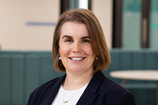 A woman with shoulder-length brown hair smiles confidently. She wears a dark blazer over a white shirt. The background features soft, modern seating and a bright environment.