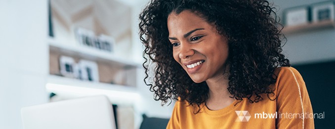 A woman with curly hair smiles while looking at a laptop screen. She is wearing an orange shirt, seated in a modern, well-lit room with soft furnishings and wall decor.