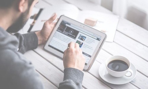 A person sitting at a wooden table interacts with a tablet, reading content while holding a stylus. A cup of coffee sits beside them, and an open magazine is visible nearby.