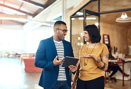 Two colleagues are engaged in a conversation, sharing a tablet. They are in a bright, modern office with casual furniture and large windows, creating a collaborative atmosphere.