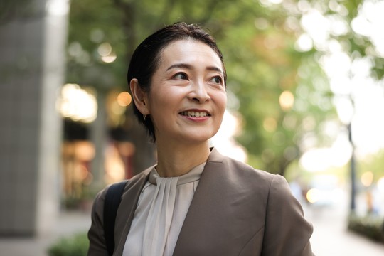 A smiling woman with short hair wears a business suit, looking upwards. She stands on a tree-lined street, with soft sunlight filtering through the leaves and a blurred city background.
