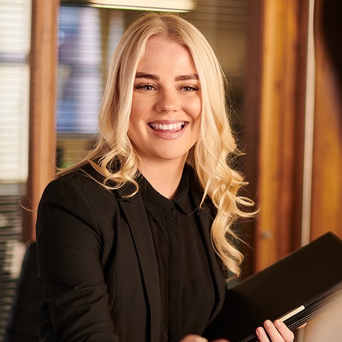 A woman with long, blonde hair smiles while holding a notebook. She is engaging with someone out of view in a well-lit office space with wooden accents and blinds.