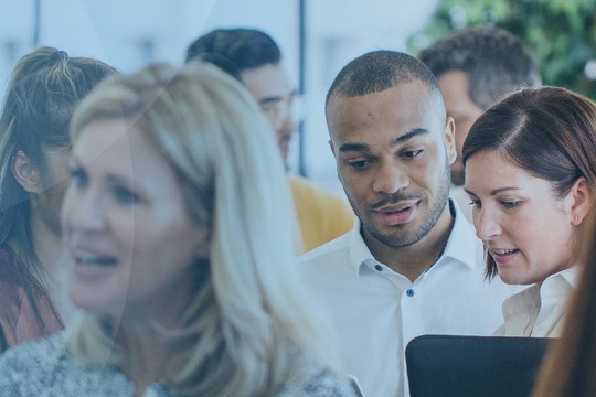 A group of professionals interacts in a bright, modern office space. Two individuals closely examine a device or document while others converse, creating a vibrant atmosphere of collaboration.