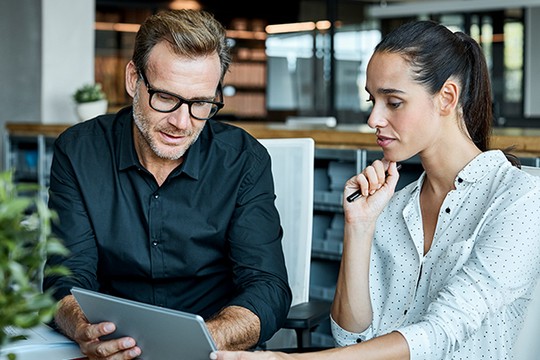 A man and a woman sit at a table, engaged in discussion while examining a tablet. The office setting features modern decor, abundant natural light, and a relaxed ambiance.