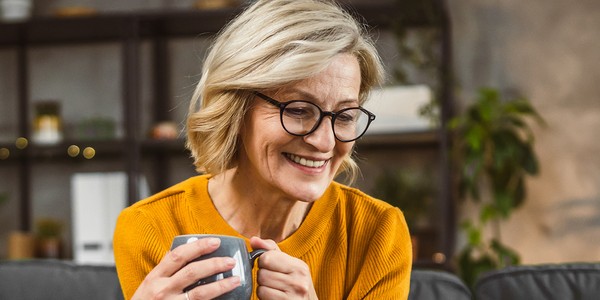 A middle-aged woman with short, wavy blonde hair smiles while holding a gray mug. She is seated on a couch in a cozy, well-decorated living room filled with plants and shelves.