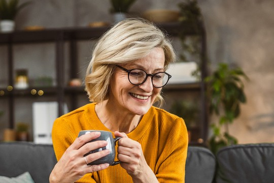 A middle-aged woman with short, wavy blonde hair smiles while holding a gray mug. She is seated on a couch in a cozy, well-decorated living room filled with plants and shelves.