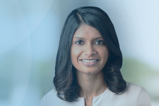 A woman with long, dark hair smiles confidently. She is dressed in light clothing against a soft blue background, suggesting a professional and inviting atmosphere.