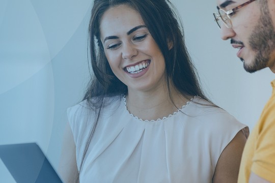 A woman with dark hair smiles while looking at a tablet held by a man in a yellow shirt. They are engaged in conversation in a bright, modern setting.