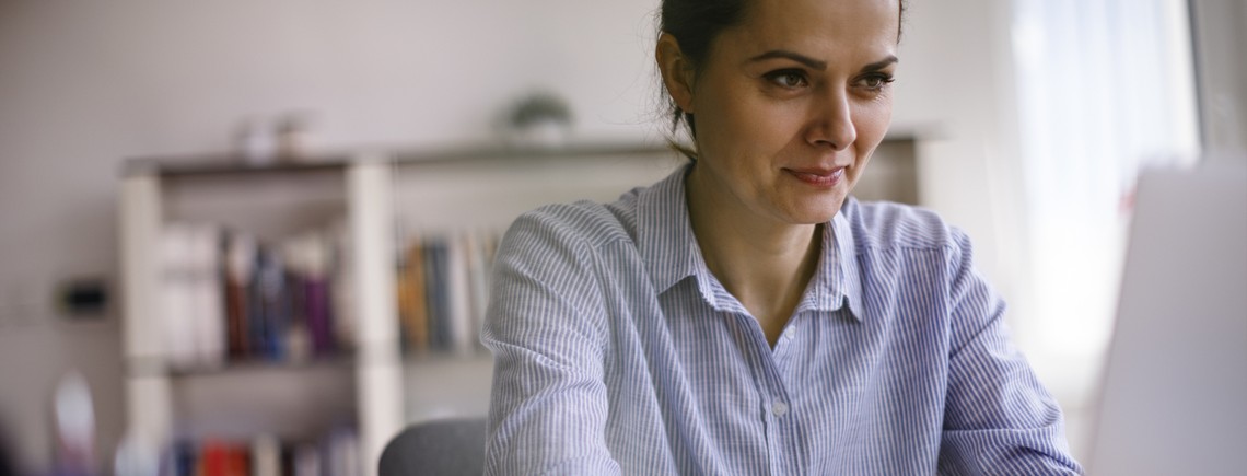 A woman in a striped shirt focuses intently on her laptop, set against a soft, well-lit background with shelves containing books, conveying a productive and calm workspace atmosphere.